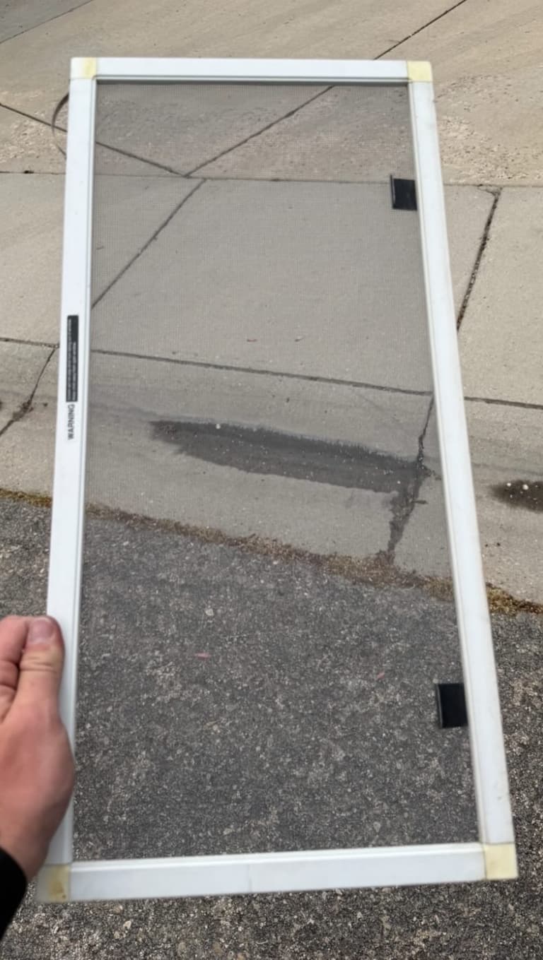 Hand holding a rectangular white-framed window screen with fine mesh over a concrete sidewalk.